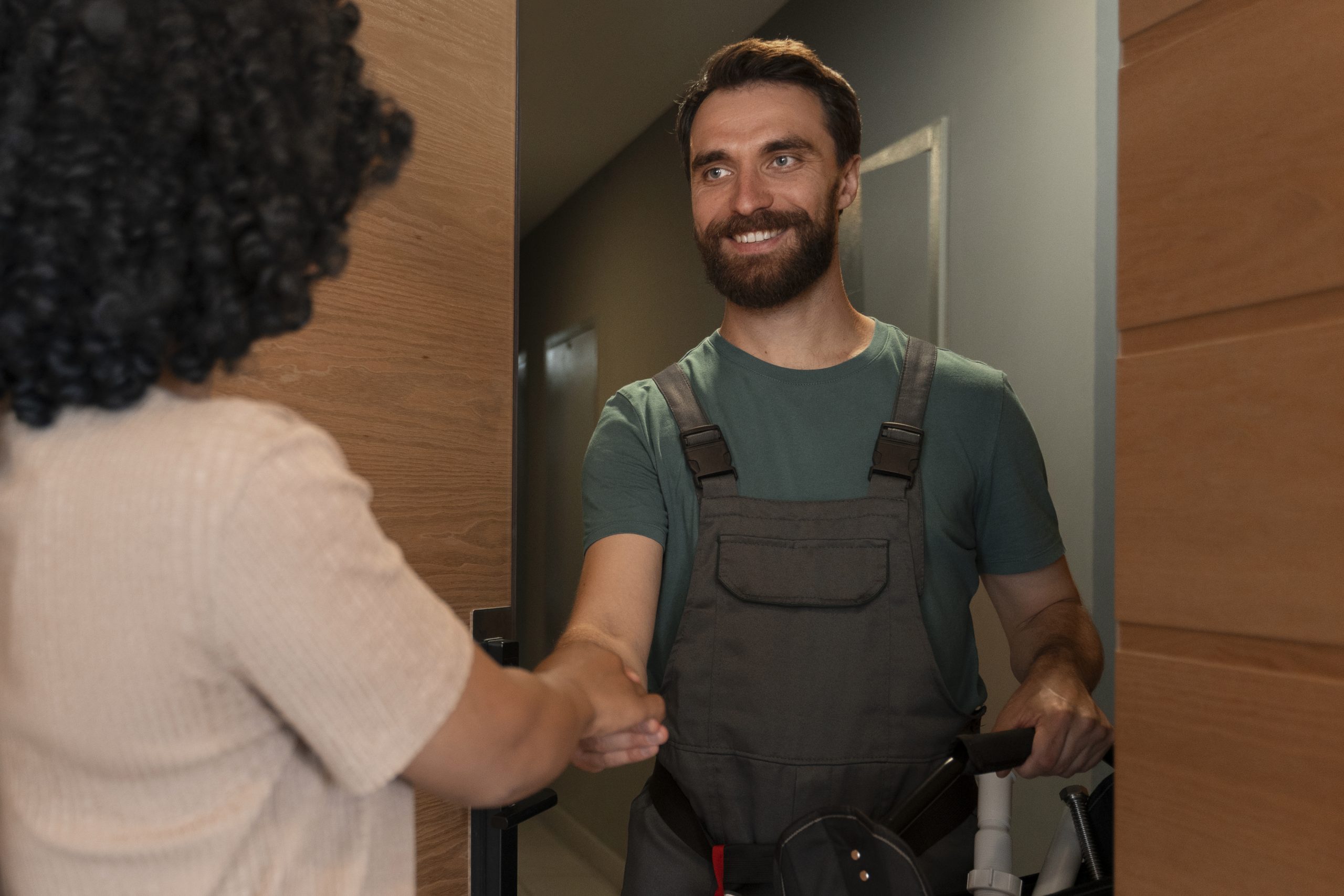 A smiling tradesman in overalls and a green t-shirt shaking hands with a customer at their front door.Retry